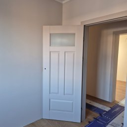 Usługi ogólnobudowlane Maciej Rowiński - Białe drzwi z szybą w odcieniach szarości, zamontowane w odnowionym wnętrzu z panelami podłogowymi i świeżo pomalowanymi ścianami. Widoczne zabezpieczenia podłogi.