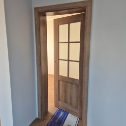Usługi ogólnobudowlane Maciej Rowiński - Otwarta, drewniana, przeszklona wewnętrzna drzwi z ościeżnicą, po montażu. Na podłodze leżą panele podłogowe w opakowaniu. Ściany pomalowane na szaro.