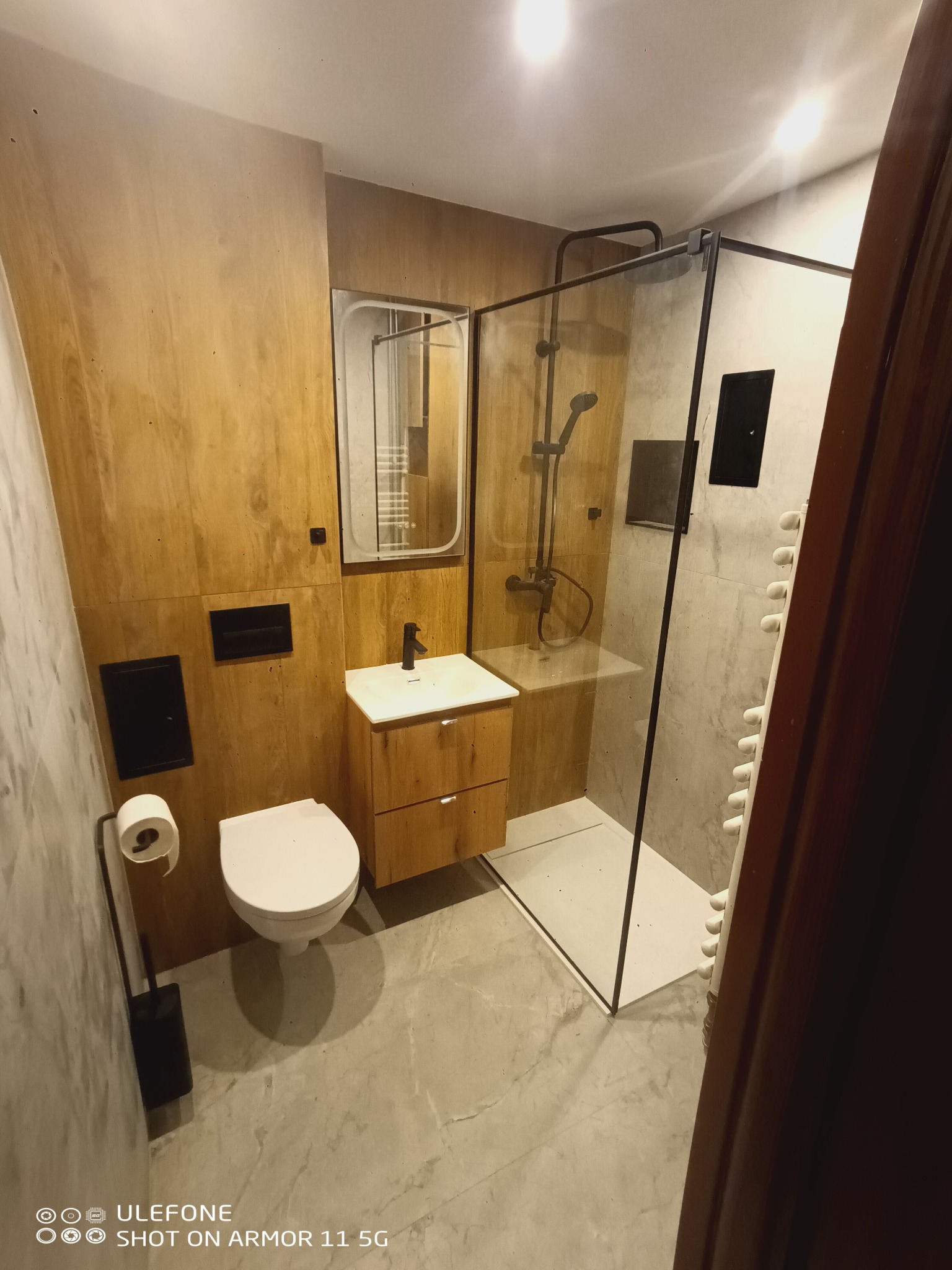 Nowoczesna łazienka z drewnianą okładziną, kabiną prysznicową typu walk-in i wiszącą toaletą. Minimalistyczny design z akcentami czerni i szarości.