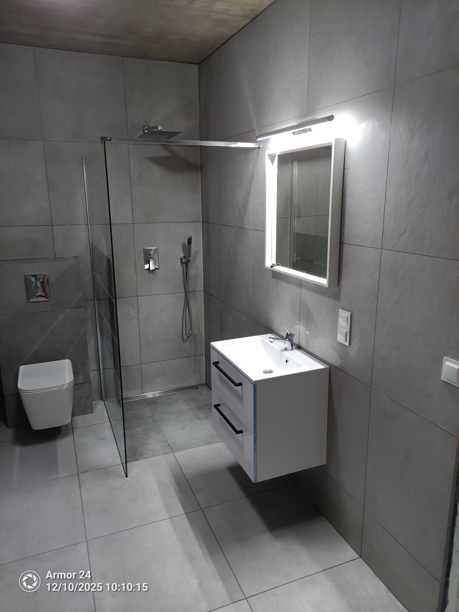 Nowoczesna, szara łazienka po remoncie: kabina prysznicowa ze szklaną ścianką, umywalka z szafką i podświetlanym lustrem. Minimalistyczny design, płytki w odcieniach szarości.