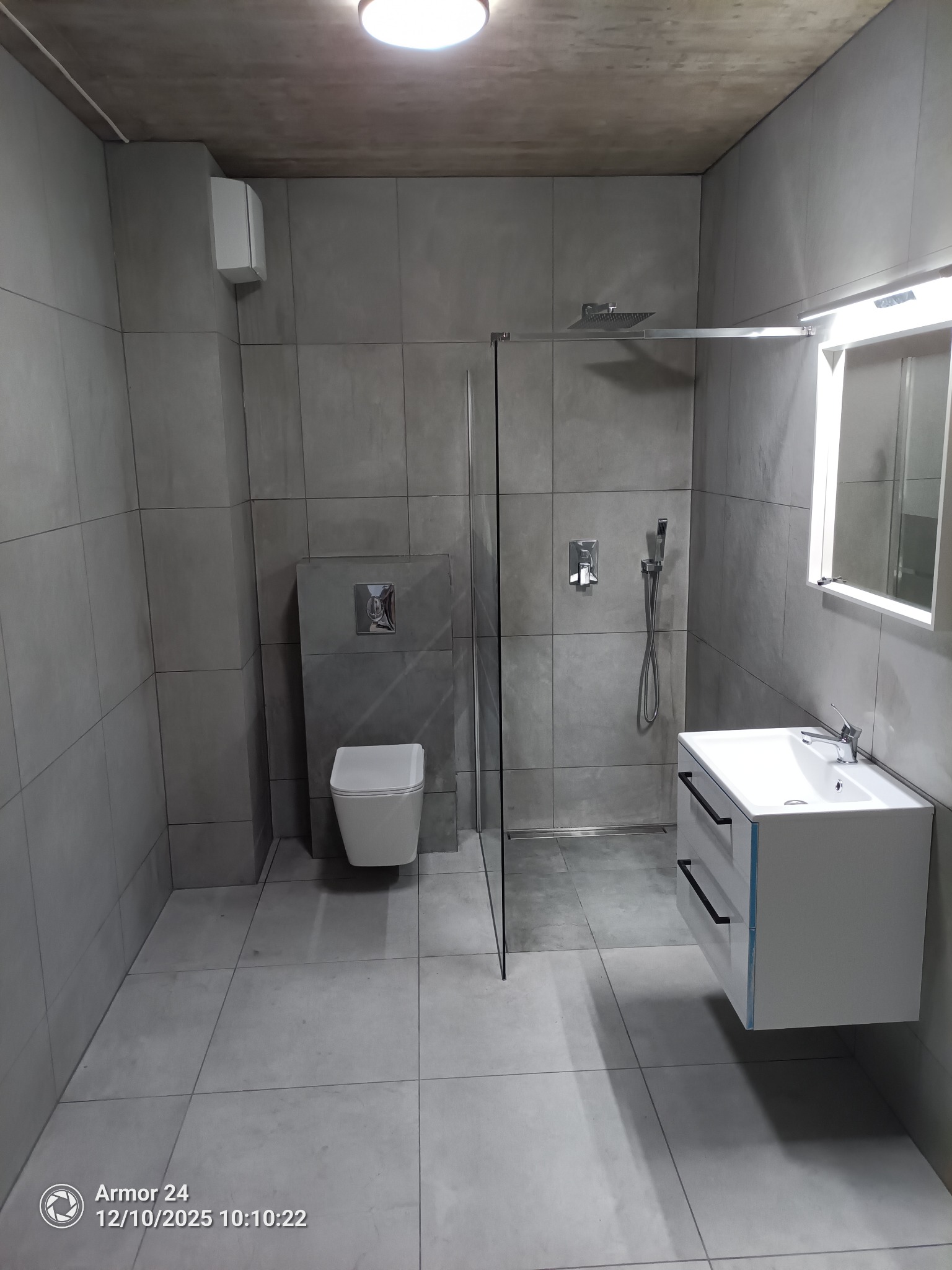 Nowoczesna łazienka w szarościach: kabina prysznicowa ze szklaną ścianką, podwieszana toaleta i umywalka z szafką. Minimalistyczny design, płytki imitujące beton.