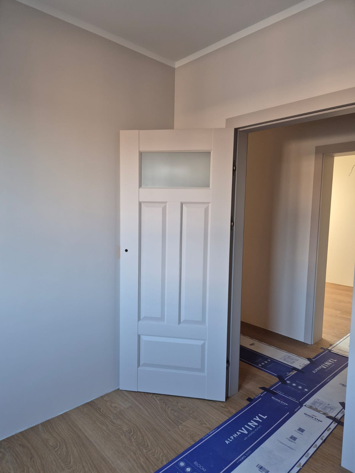 Białe drzwi z szybą w odcieniach szarości, zamontowane w odnowionym wnętrzu z panelami podłogowymi i świeżo pomalowanymi ścianami. Widoczne zabezpieczenia podłogi.