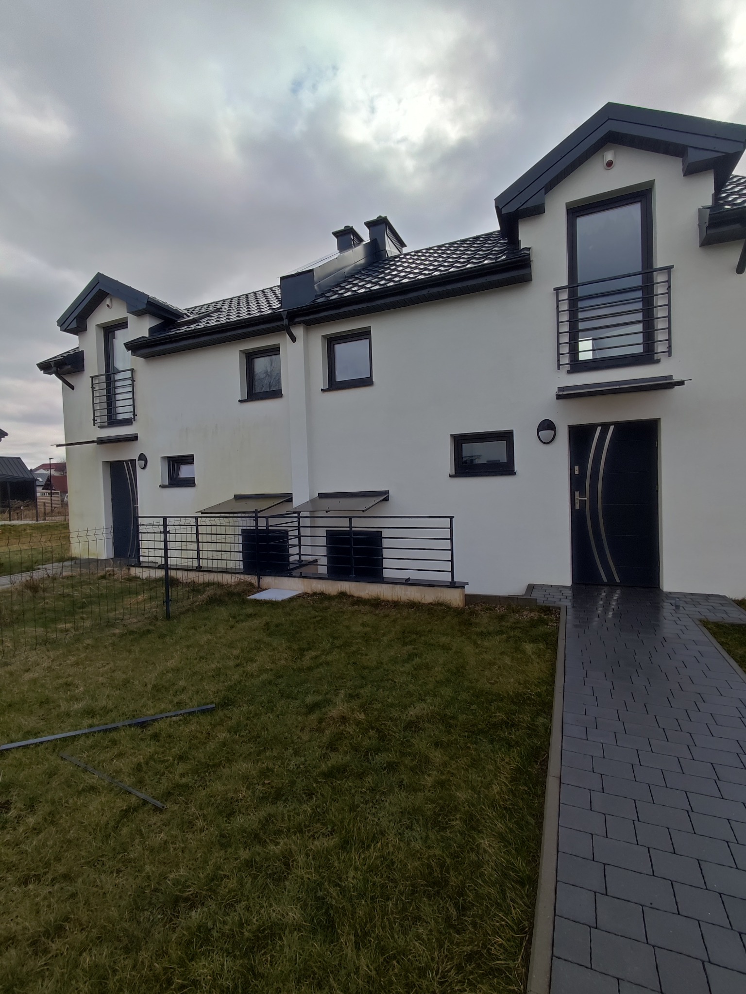 Elewacja szeregowca w jasnym kolorze z ciemnymi ramami okiennymi i dachem, widok z poziomu trawnika w pochmurny dzień.