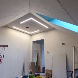 RENOWEJT Przemysław Saniewski - Poddasze w trakcie prac wykończeniowych: zamontowane płyty karton-gips, widoczne profile LED w suficie, podnośnik do płyt oraz materiały budowlane.