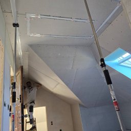 RENOWEJT Przemysław Saniewski - Pracownik na drabinie montuje płytę kartonowo-gipsową na poddaszu, widoczne metalowe profile pod konstrukcję i okno dachowe.