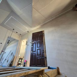 RENOWEJT Przemysław Saniewski - Wnętrze w trakcie prac wykończeniowych: ściany ze świeżo nałożoną gładzią, widoczne płyty kartonowo-gipsowe na suficie z zamontowanymi profilami oświetleniowymi, drewniane rusztowanie i drzwi...
