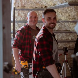 RENOWEJT Przemysław Saniewski - Dwóch uśmiechniętych mężczyzn w flanelowych koszulach, z narzędziami przy pasie, w trakcie ocieplania poddasza pianką PUR, widoczne metalowe profile i warstwa izolacyjna.