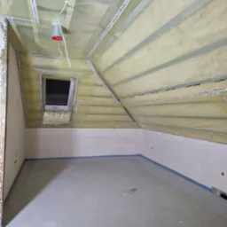 Poddasze w trakcie ocieplania, widoczne folie paroizolacyjne z taśmami uszczelniającymi, metalowe profile, okno dachowe oraz przygotowane ściany do dalszych prac wykończeniowych.