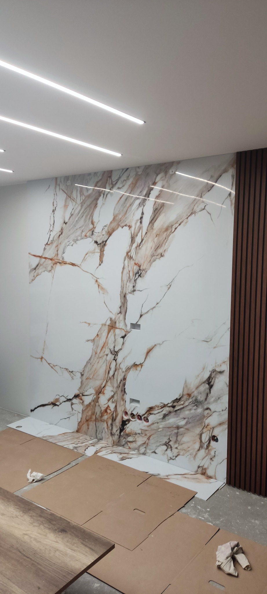 Wykończenie ściany w Łomży z użyciem płyty imitującej marmur, z widocznymi gniazdkami elektrycznymi i drewnianymi panelami dekoracyjnymi po prawej stronie, podłoga zabezpieczona kartonem.