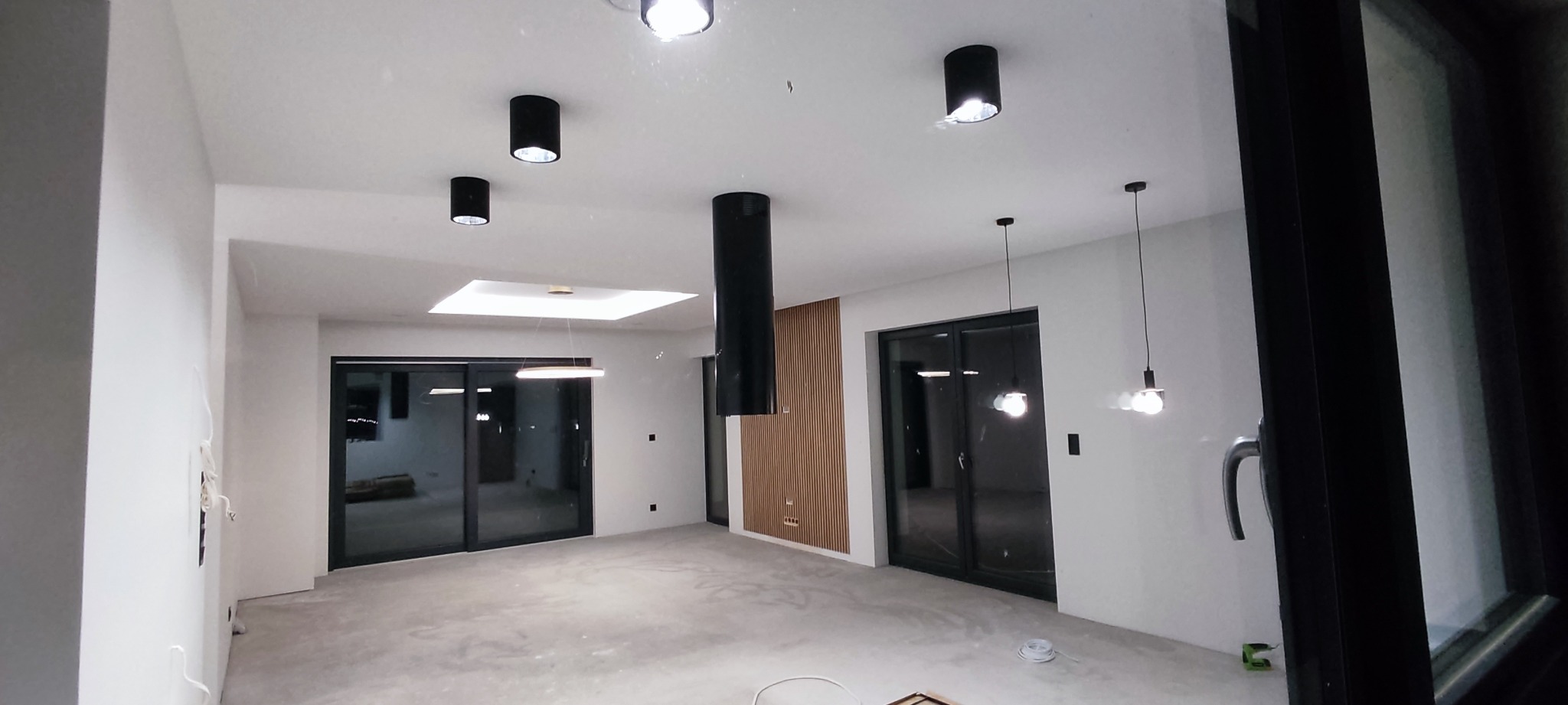 Wnętrze domu w trakcie wykańczania: gładkie, białe ściany po szpachlowaniu, surowa betonowa podłoga, zamontowane czarne lampy sufitowe i przesuwne drzwi balkonowe z czarnymi ramami.