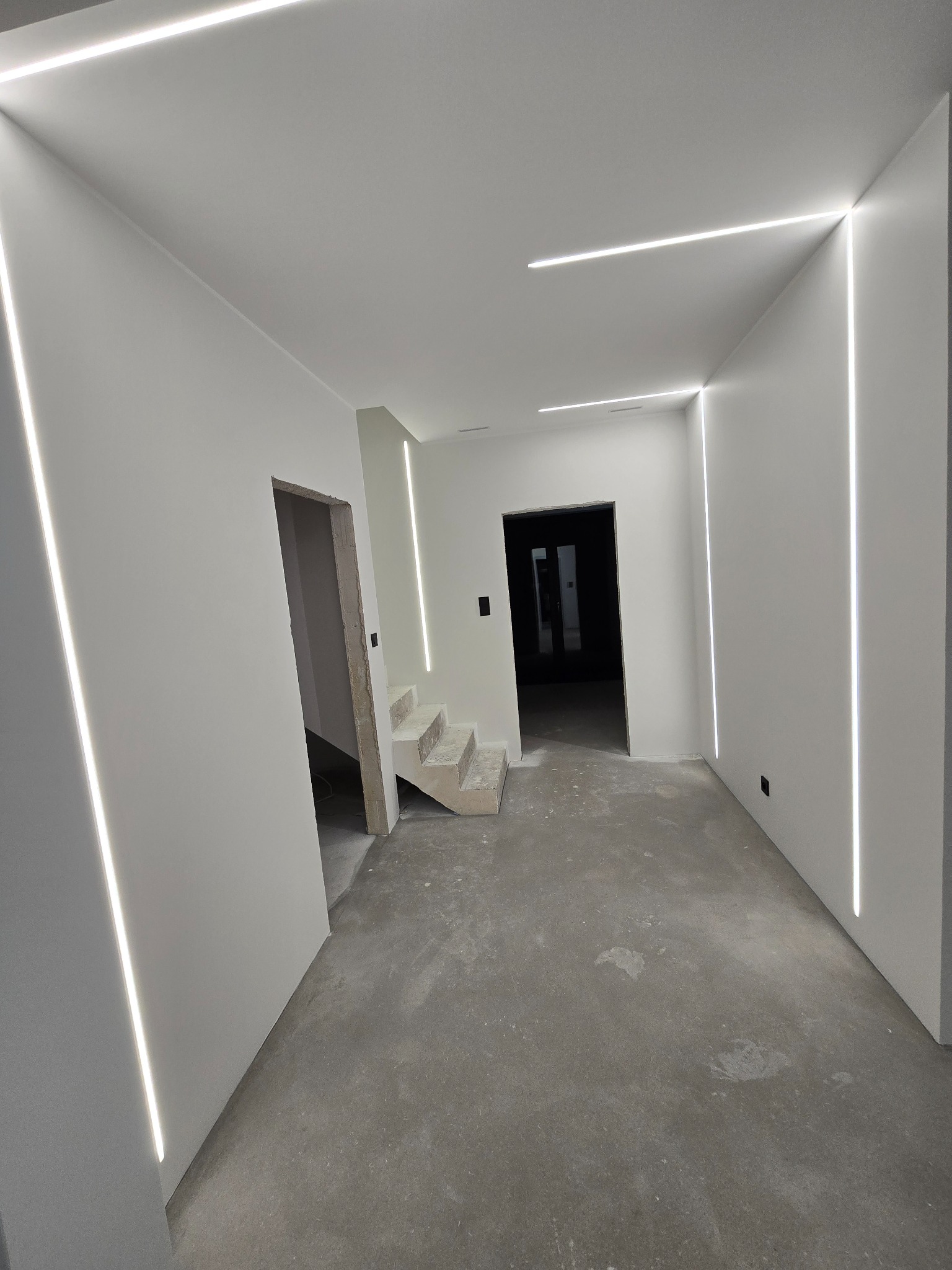 Wnętrze w trakcie prac wykończeniowych z surową betonową podłogą, białymi ścianami z wbudowanym oświetleniem LED w pionowe i poziome linie, widoczne schody w stanie surowym oraz otwory drzwiowe bez...