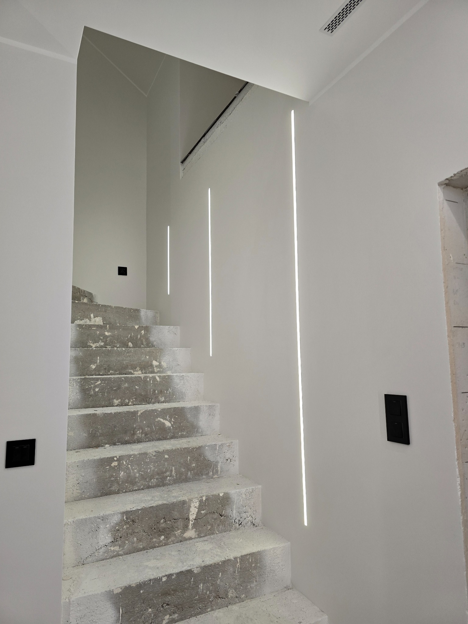 Surowe betonowe schody w trakcie prac wykończeniowych, z minimalistycznym oświetleniem LED wpuszczonym w białą ścianę i czarnymi włącznikami.