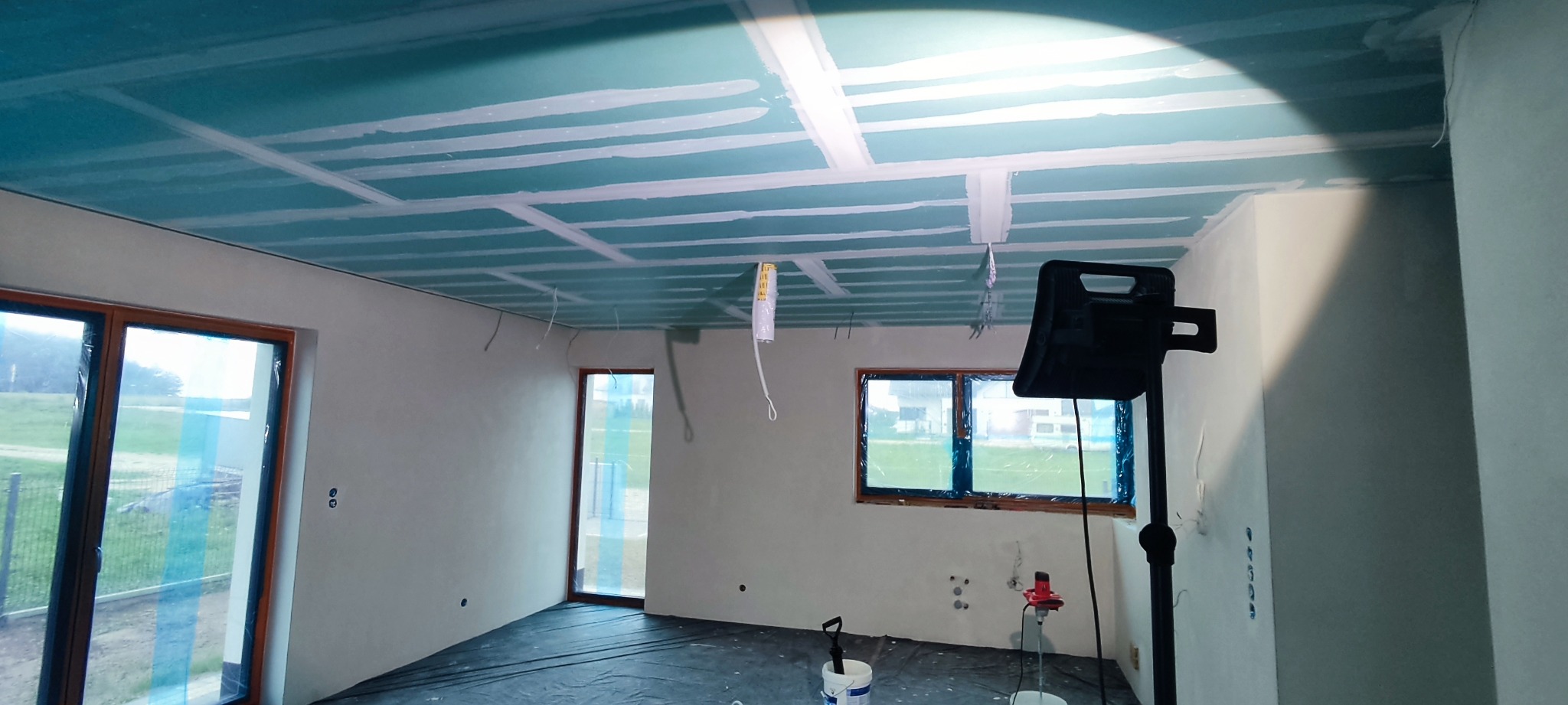 Wnętrze w trakcie prac wykończeniowych: sufit z płyt kartonowo-gipsowych z widocznymi łączeniami, przygotowany do dalszej obróbki, ściany po szpachlowaniu, podłoga zabezpieczona folią, na środku...