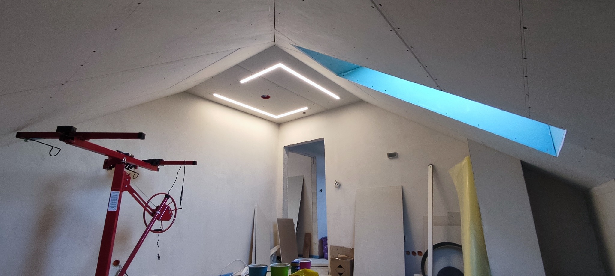 Poddasze w trakcie prac wykończeniowych: zamontowane płyty karton-gips, widoczne profile LED w suficie, podnośnik do płyt oraz materiały budowlane.