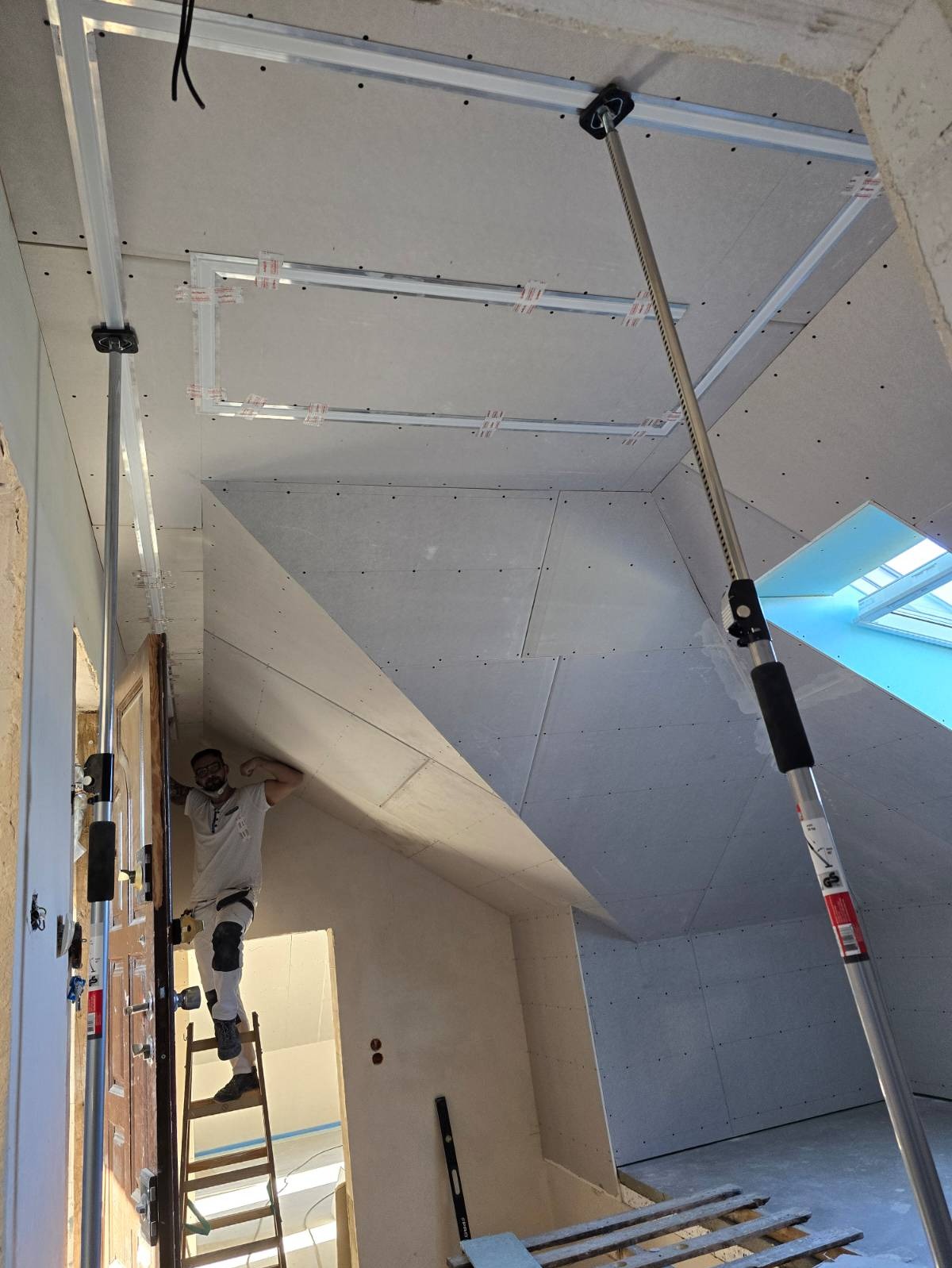 Pracownik na drabinie montuje płytę kartonowo-gipsową na poddaszu, widoczne metalowe profile pod konstrukcję i okno dachowe.
