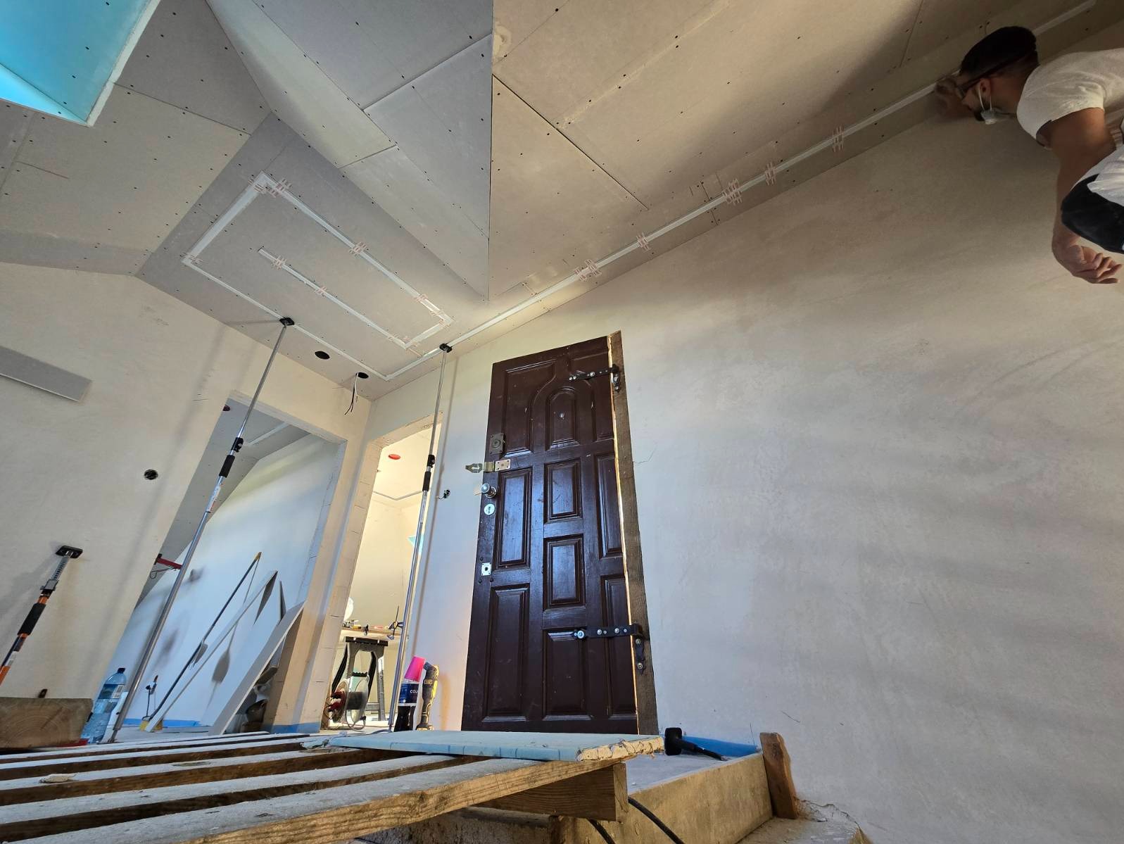 Wnętrze w trakcie prac wykończeniowych: ściany ze świeżo nałożoną gładzią, widoczne płyty kartonowo-gipsowe na suficie z zamontowanymi profilami oświetleniowymi, drewniane rusztowanie i drzwi...