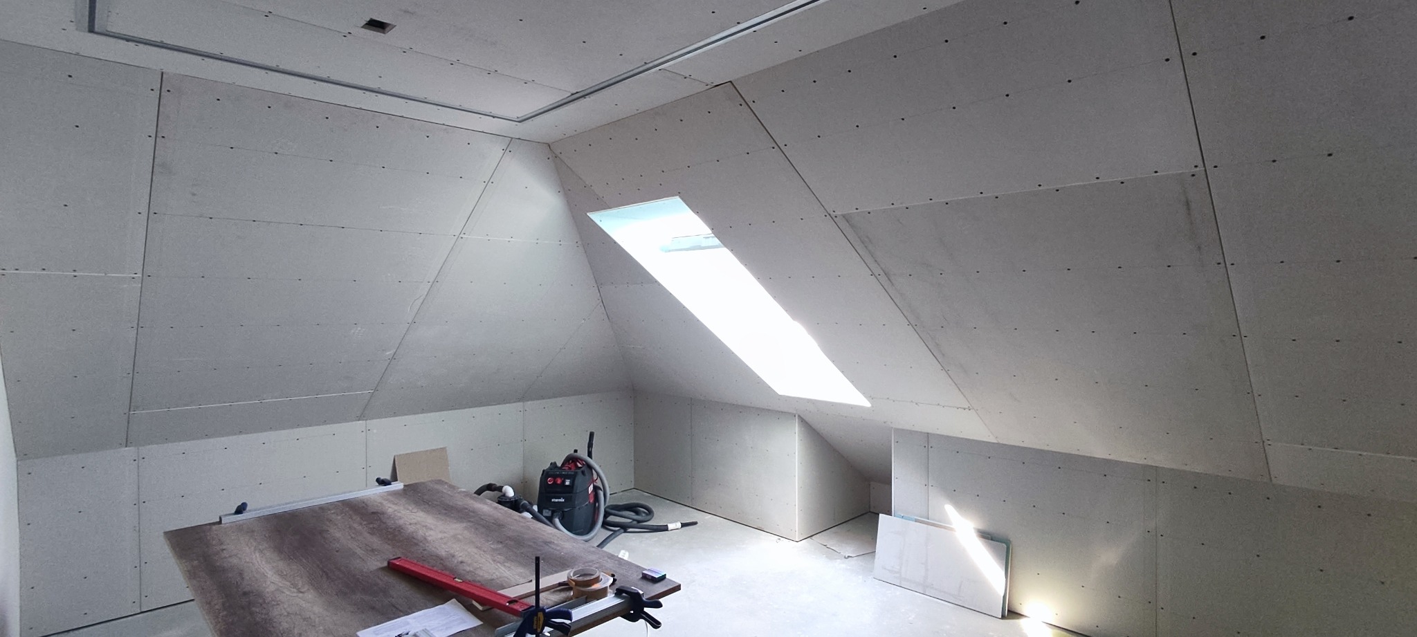 Poddasze z oknem dachowym wykończone płytami kartonowo-gipsowymi, widoczny stół roboczy z narzędziami i odkurzacz przemysłowy.