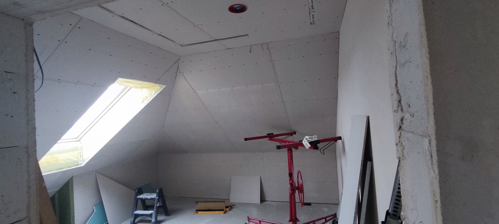 Poddasze z zamontowaną płytą karton gips z widocznym oknem dachowym, drabinką i podnośnikiem do płyt.