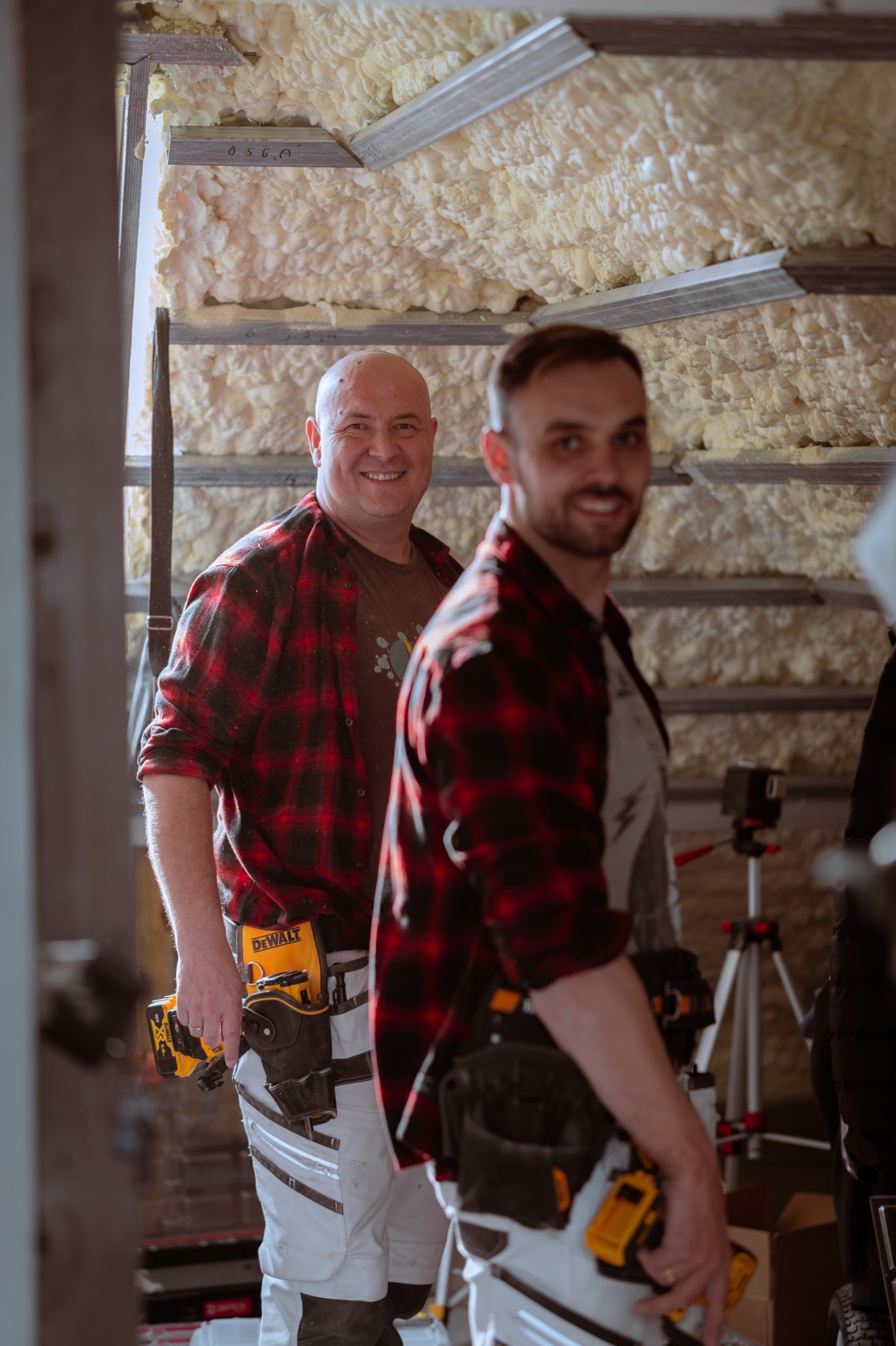 Dwóch uśmiechniętych mężczyzn w flanelowych koszulach, z narzędziami przy pasie, w trakcie ocieplania poddasza pianką PUR, widoczne metalowe profile i warstwa izolacyjna.