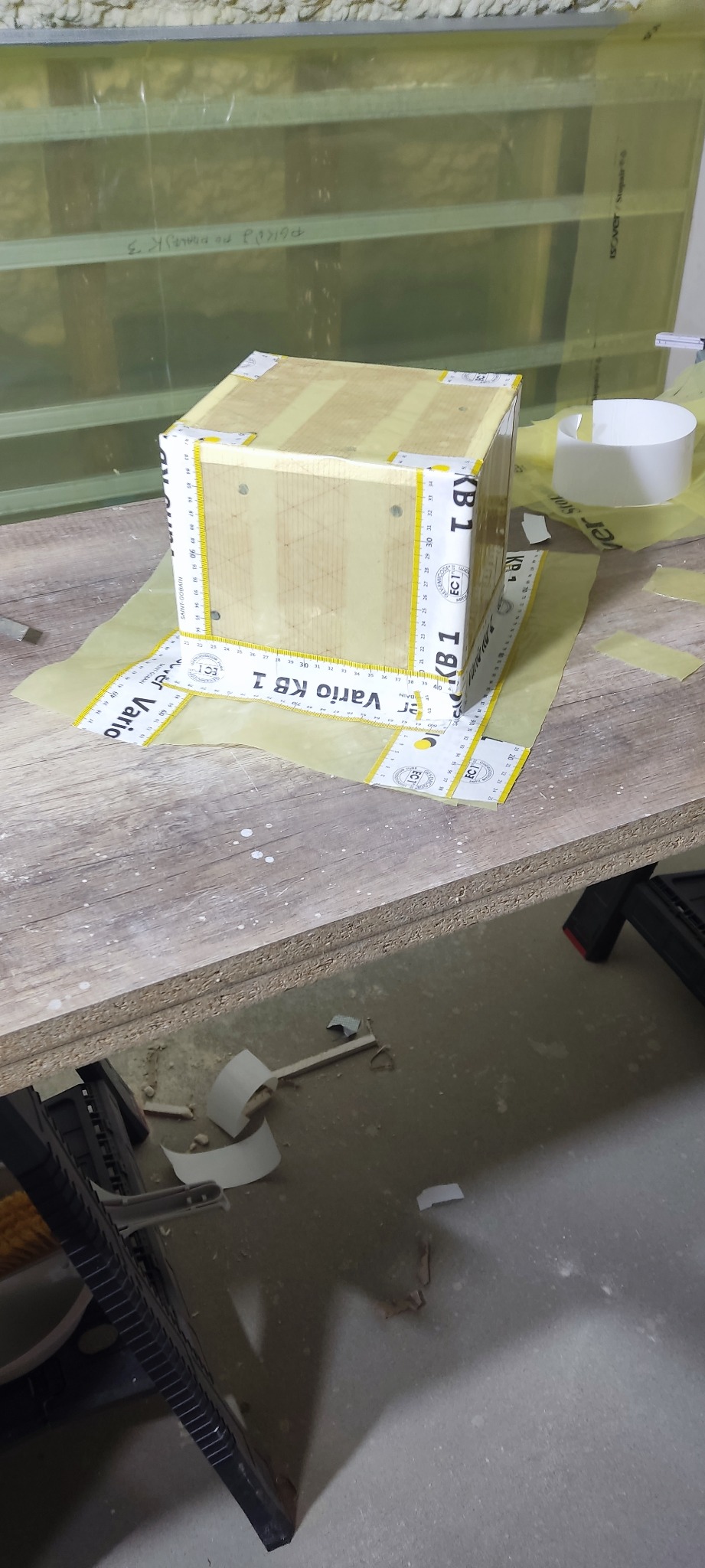 Kartonowy sześcian z płyt gipsowo-kartonowych, zabezpieczony taśmą mierniczą Vario KB 1, na stole roboczym w trakcie prac wykończeniowych.