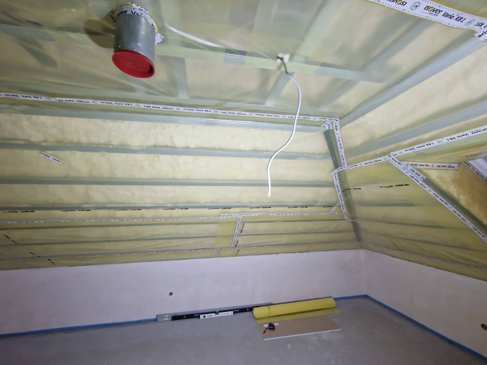 Poddasze w trakcie ocieplania: widoczne wełna mineralna, folia paroizolacyjna, profile metalowe, taśmy uszczelniające, przewód elektryczny, poziomica, rolka folii i płyta karton-gips na podłodze.