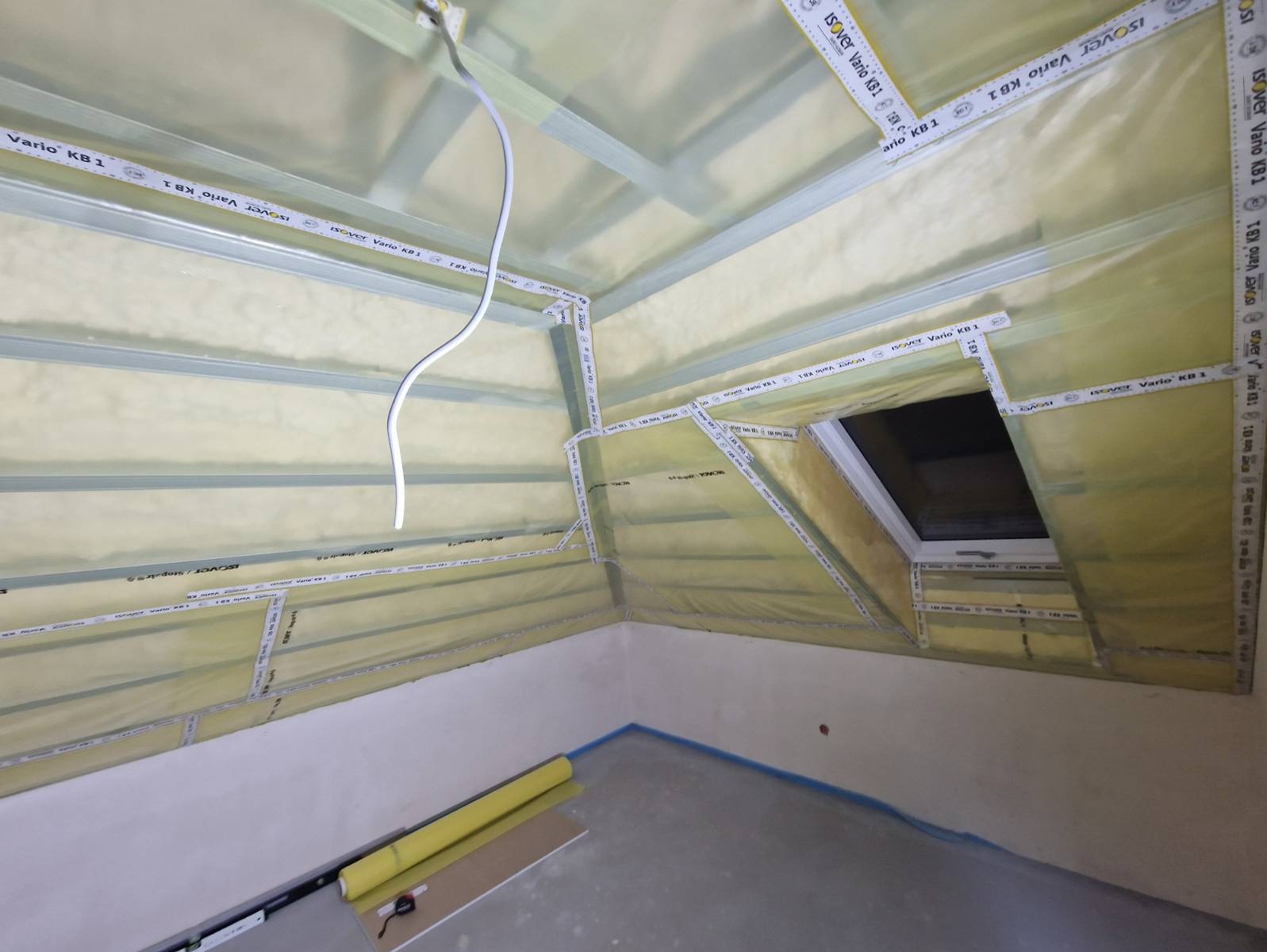 Poddasze w trakcie izolacji z widoczną folią paroizolacyjną Isover Vario KB 1, oknem dachowym i elementami konstrukcji szkieletowej, z przewodem elektrycznym zwisającym z sufitu.