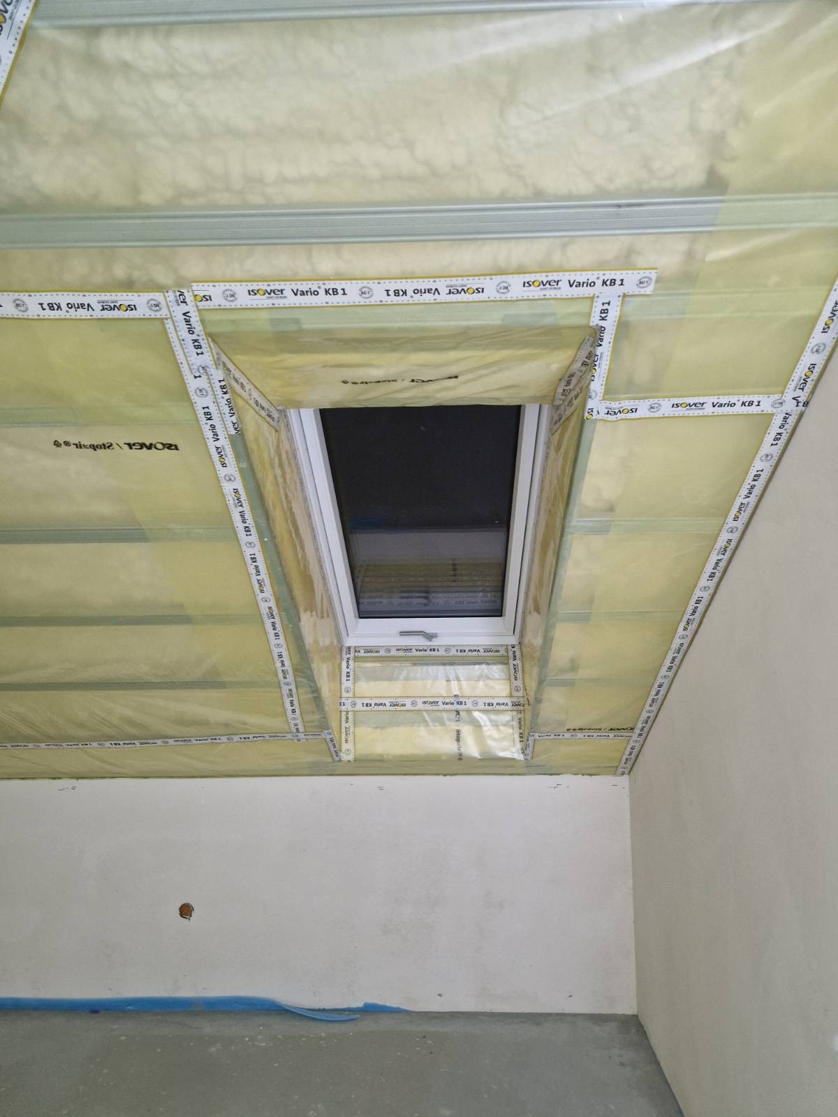 Okno dachowe w trakcie montażu ocieplenia poddasza, widoczne folie paroizolacyjne Isover Vario KB1, metalowy stelaż i surowa ściana.