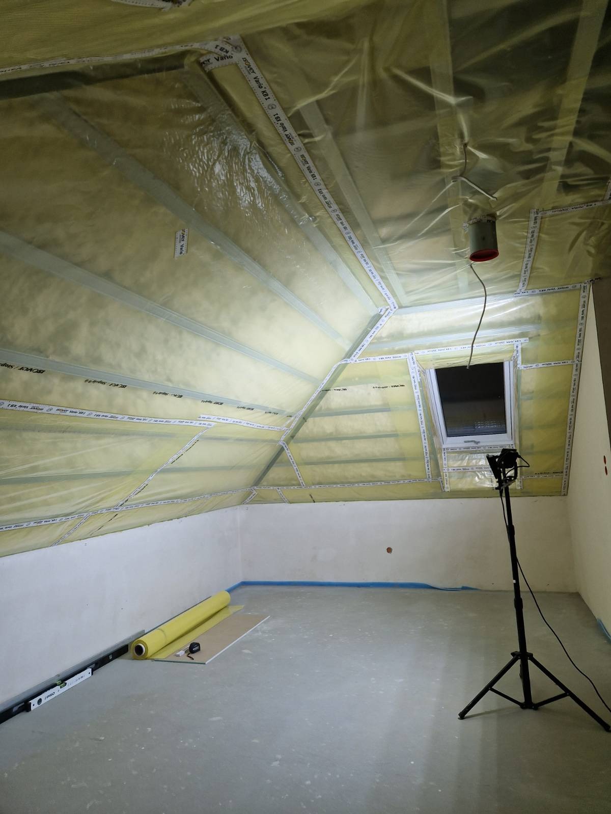 Poddasze w trakcie ocieplania: widoczne wełna mineralna, folia paroizolacyjna z nadrukiem, metalowe profile, okno dachowe oraz rozłożony na podłodze rulon folii i poziomica przy ścianie.