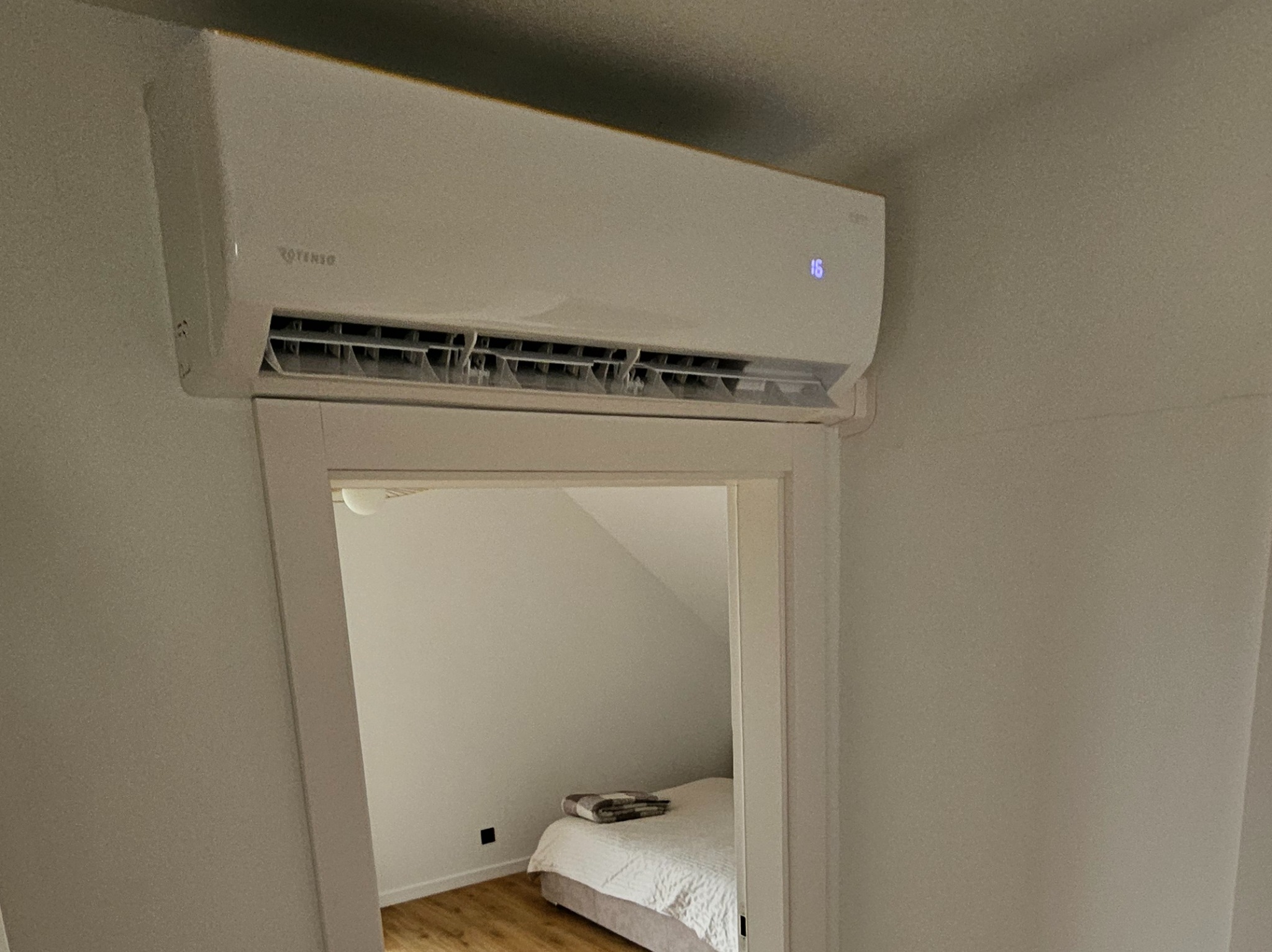 Biały klimatyzator Rotenso zamontowany nad drzwiami w pokoju na poddaszu, widok na łóżko i skos dachu. Nowoczesny design, minimalistyczne wnętrze.