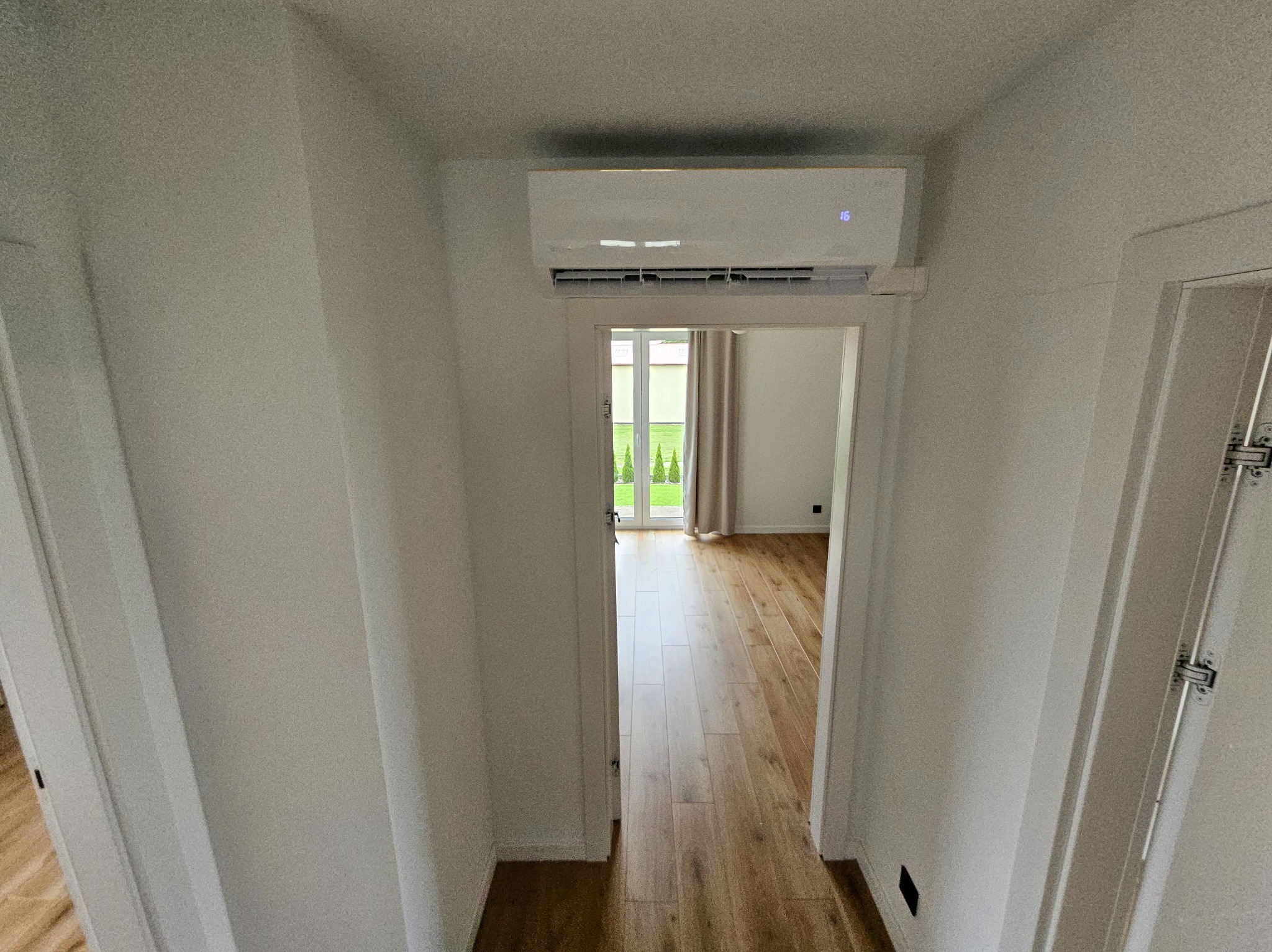 Biała klimatyzacja ścienna w jasnym wnętrzu domu. Widok na korytarz z parkietem i drzwi balkonowe. Minimalistyczny design, nowoczesne rozwiązanie klimatyzacyjne.