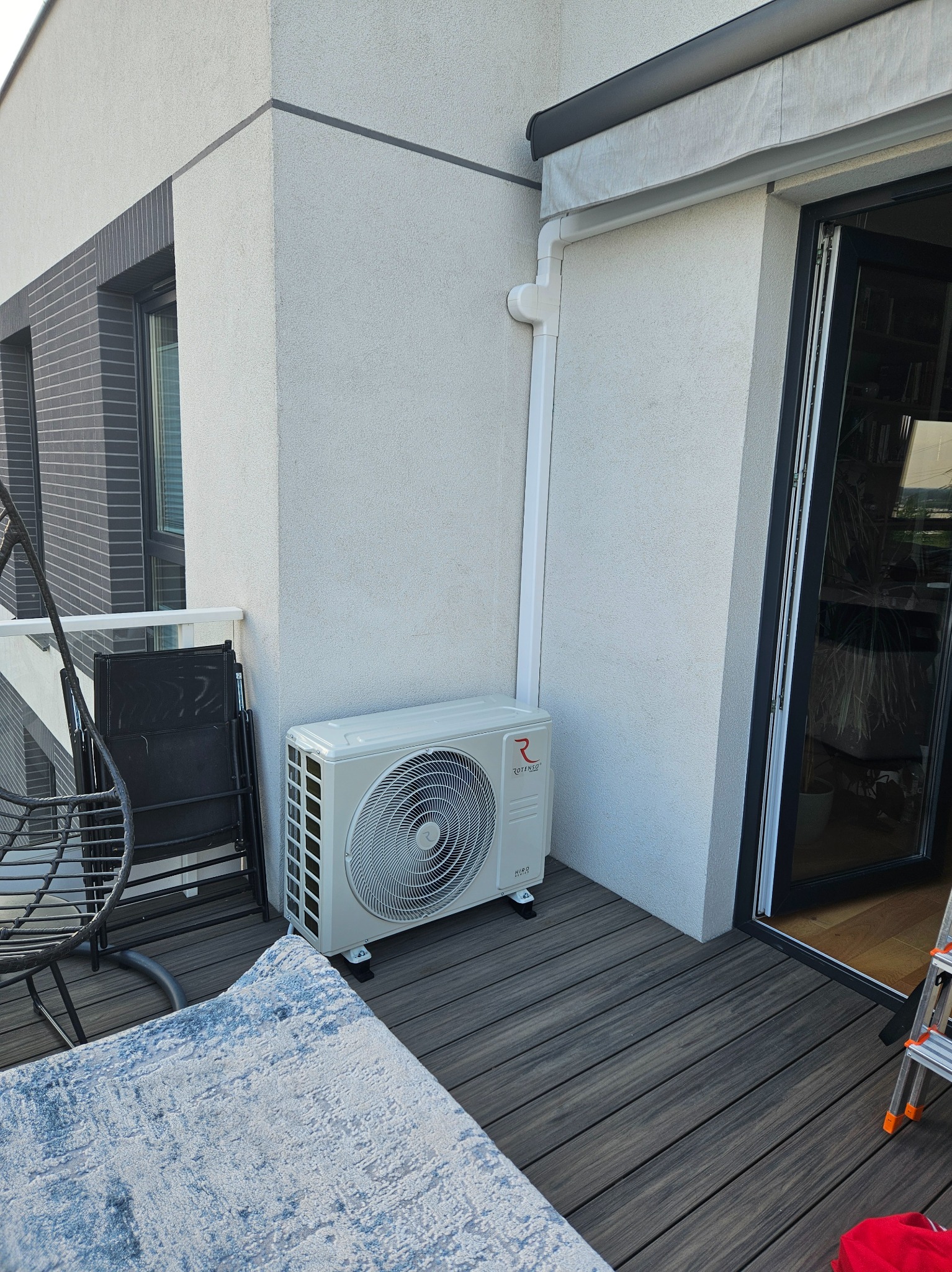 Zamontowany klimatyzator Rotenso na balkonie nowoczesnego budynku, widoczne elementy instalacji, krzesła i drabina.