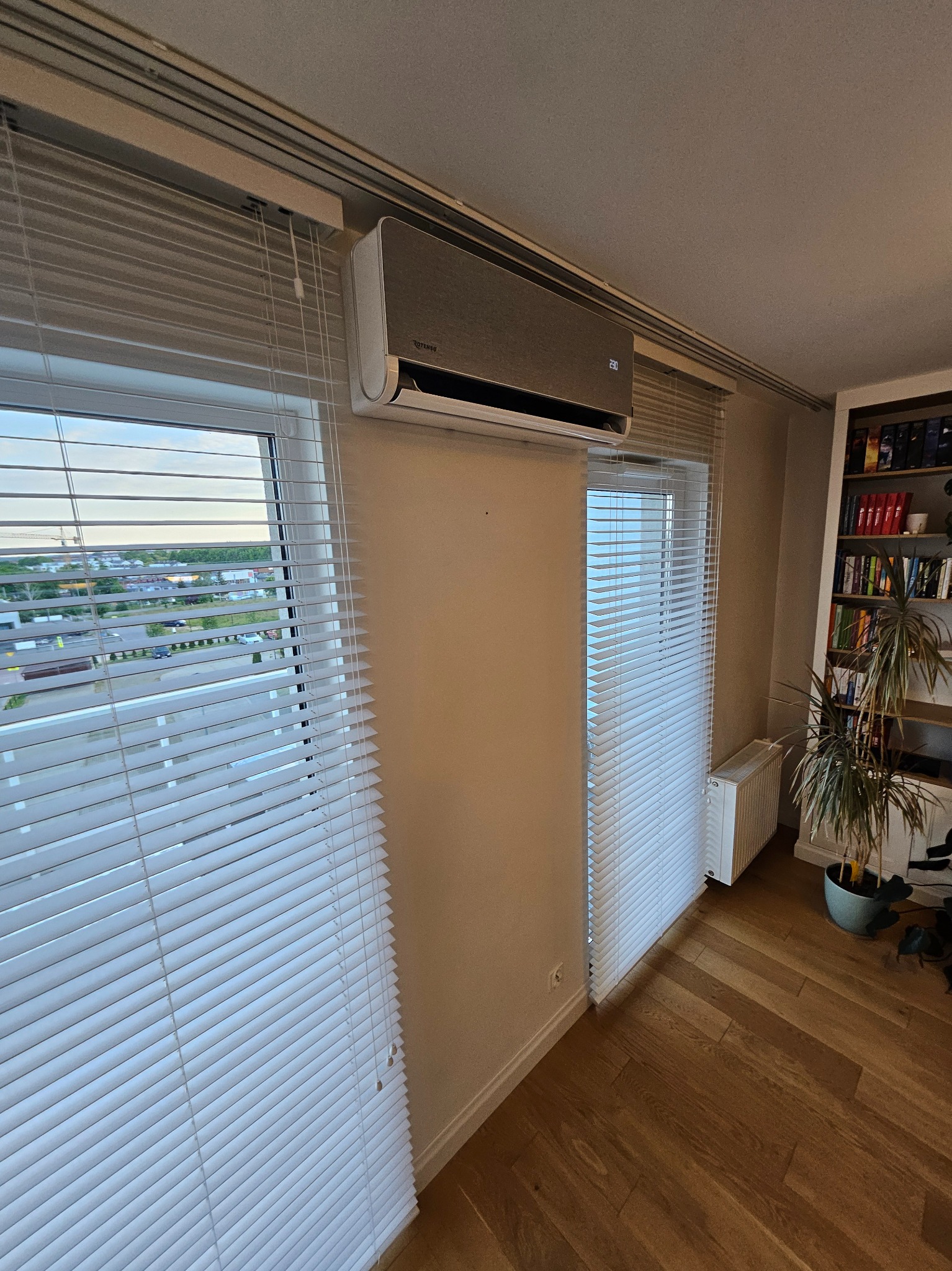 Szary klimatyzator Rotenso zamontowany nad oknem z żaluzjami w jasnym pokoju. Widoczny wyświetlacz temperatury. Na podłodze parkiet i roślina doniczkowa.