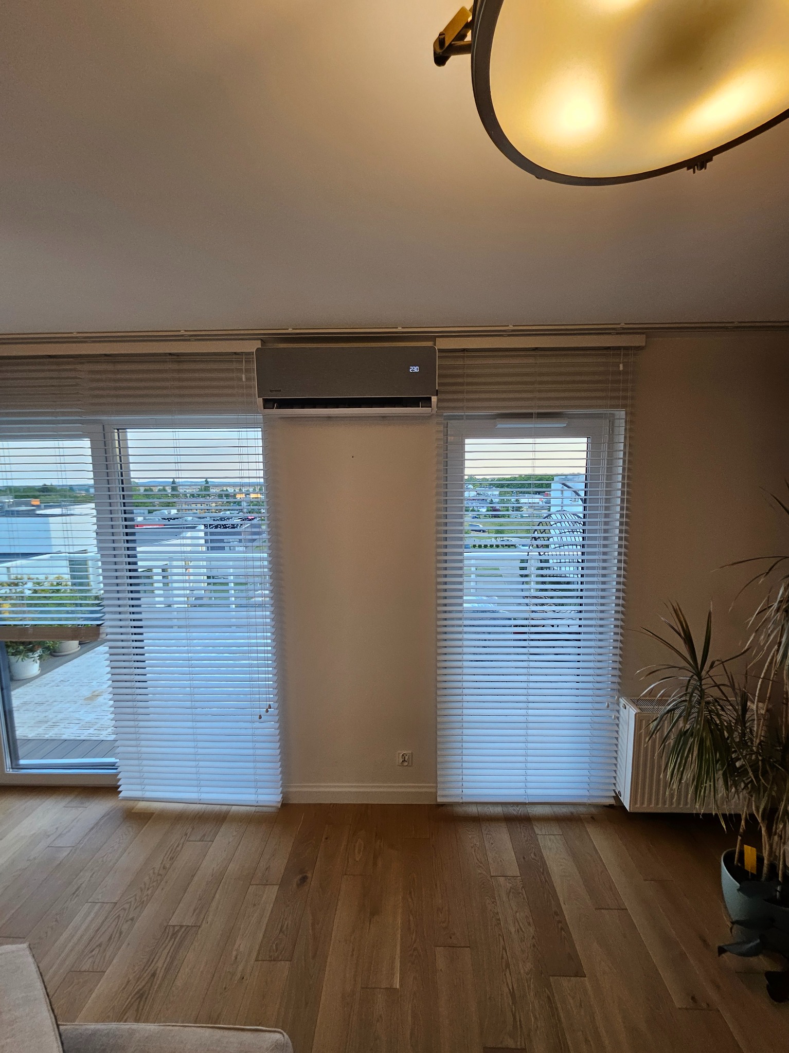 Szary klimatyzator ścienny zamontowany nad oknem z żaluzjami w jasnym wnętrzu, z widokiem na miasto. Widoczny parkiet i roślina doniczkowa.