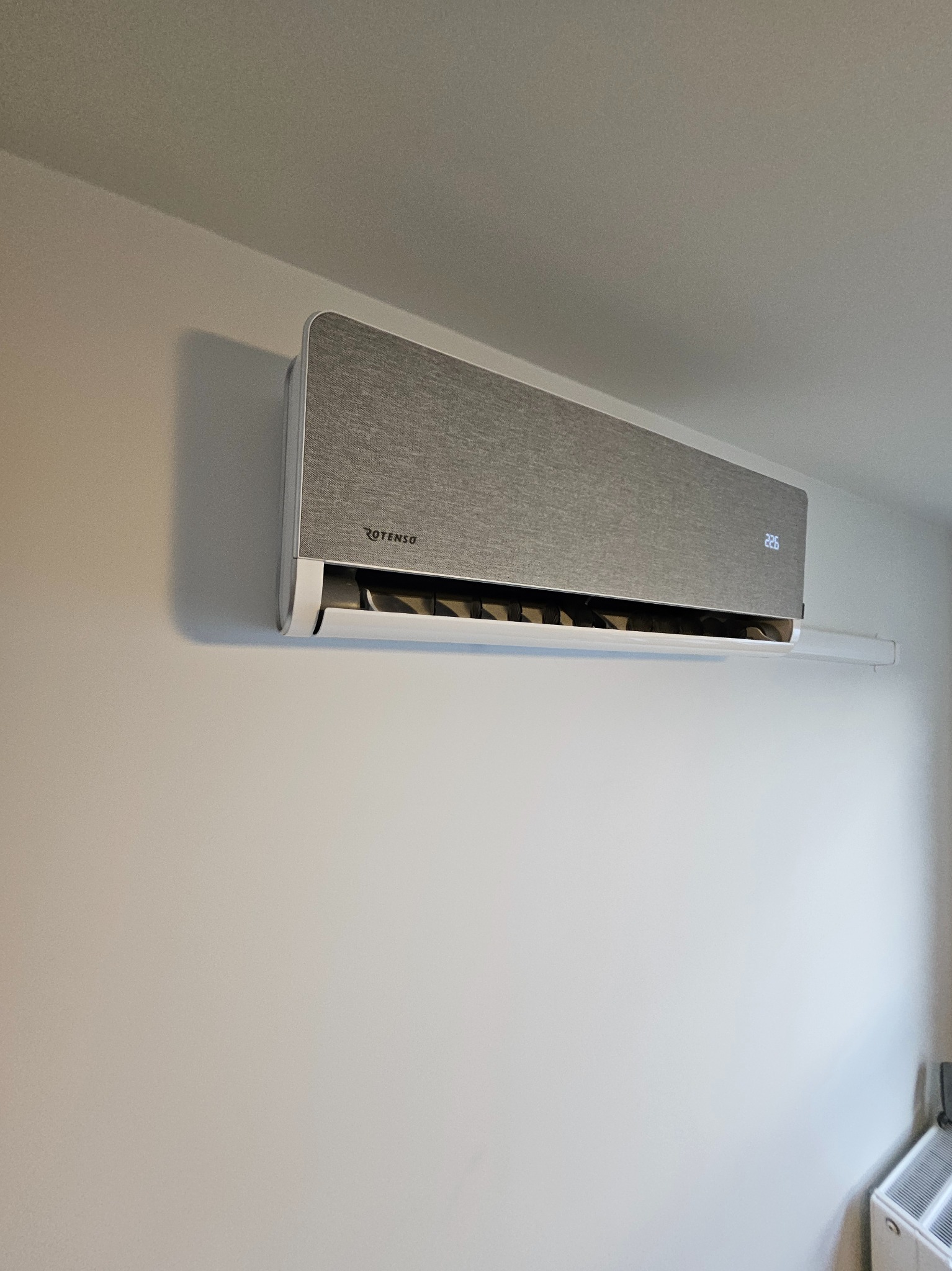 Szary klimatyzator Rotenso zamontowany na białej ścianie. Widoczny wyświetlacz temperatury 22.6°C. Minimalistyczny design w nowoczesnym wnętrzu.