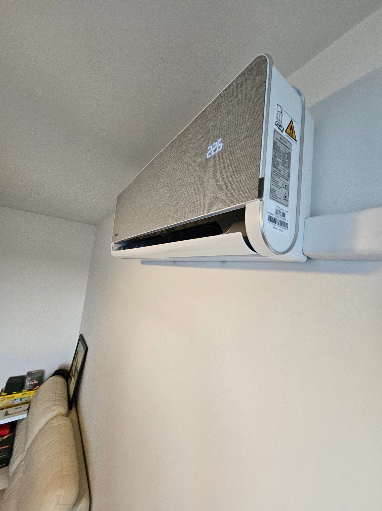 Nowoczesny klimatyzator ścienny w jasnym wnętrzu. Widoczny panel z wyświetlaczem temperatury i etykietami informacyjnymi. Minimalistyczny design.