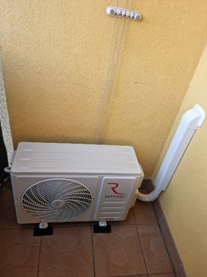 Zamontowana, biała jednostka zewnętrzna klimatyzacji Rotenso z odprowadzeniem skroplin na balkonie z brązową podłogą i żółtą ścianą. Widok z boku.