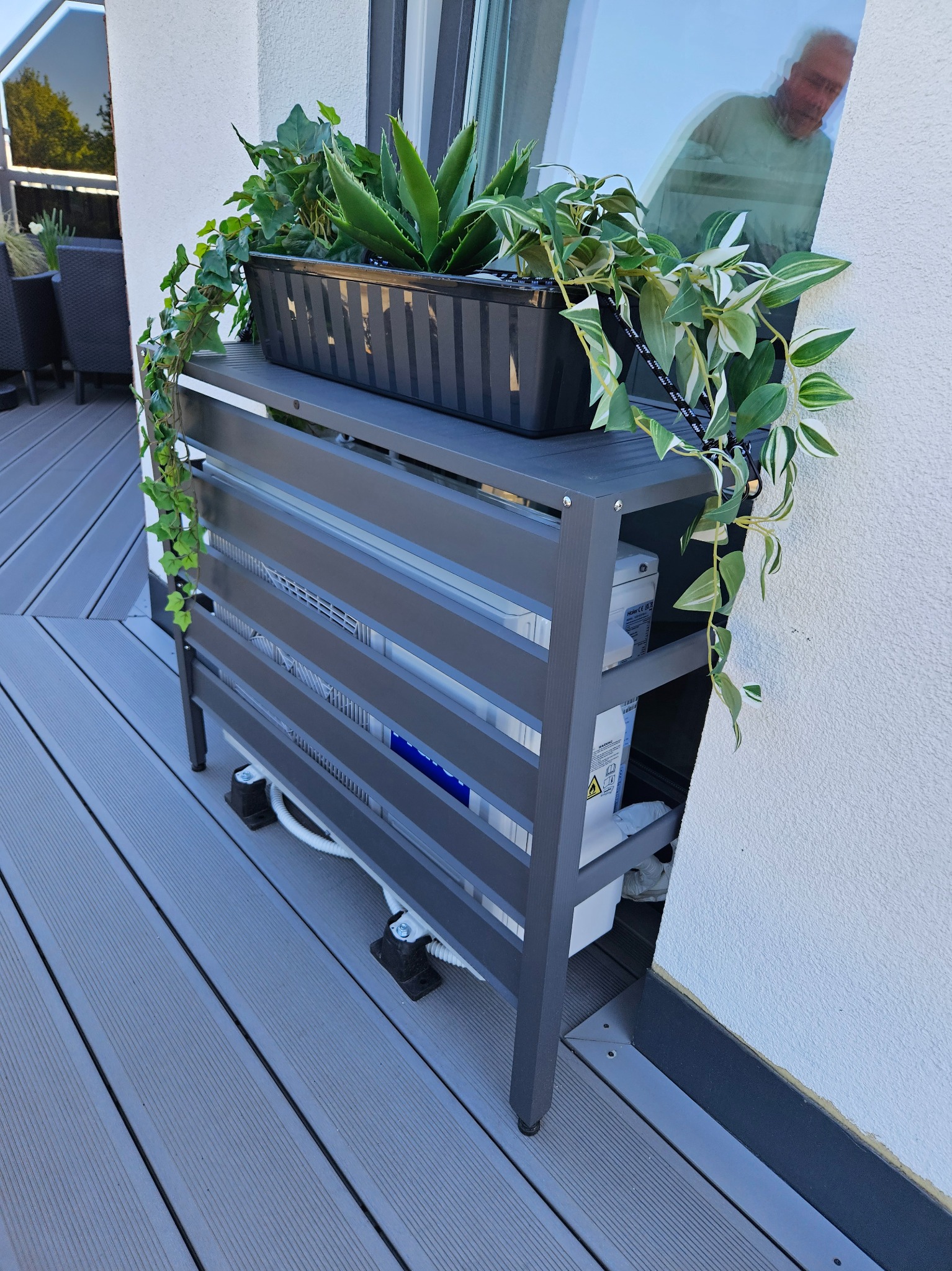 Klimatyzator na balkonie, ukryty w szarej, ażurowej obudowie z donicą na górze, z której zwisają zielone rośliny. W tle widoczne odbicie osoby w szybie.