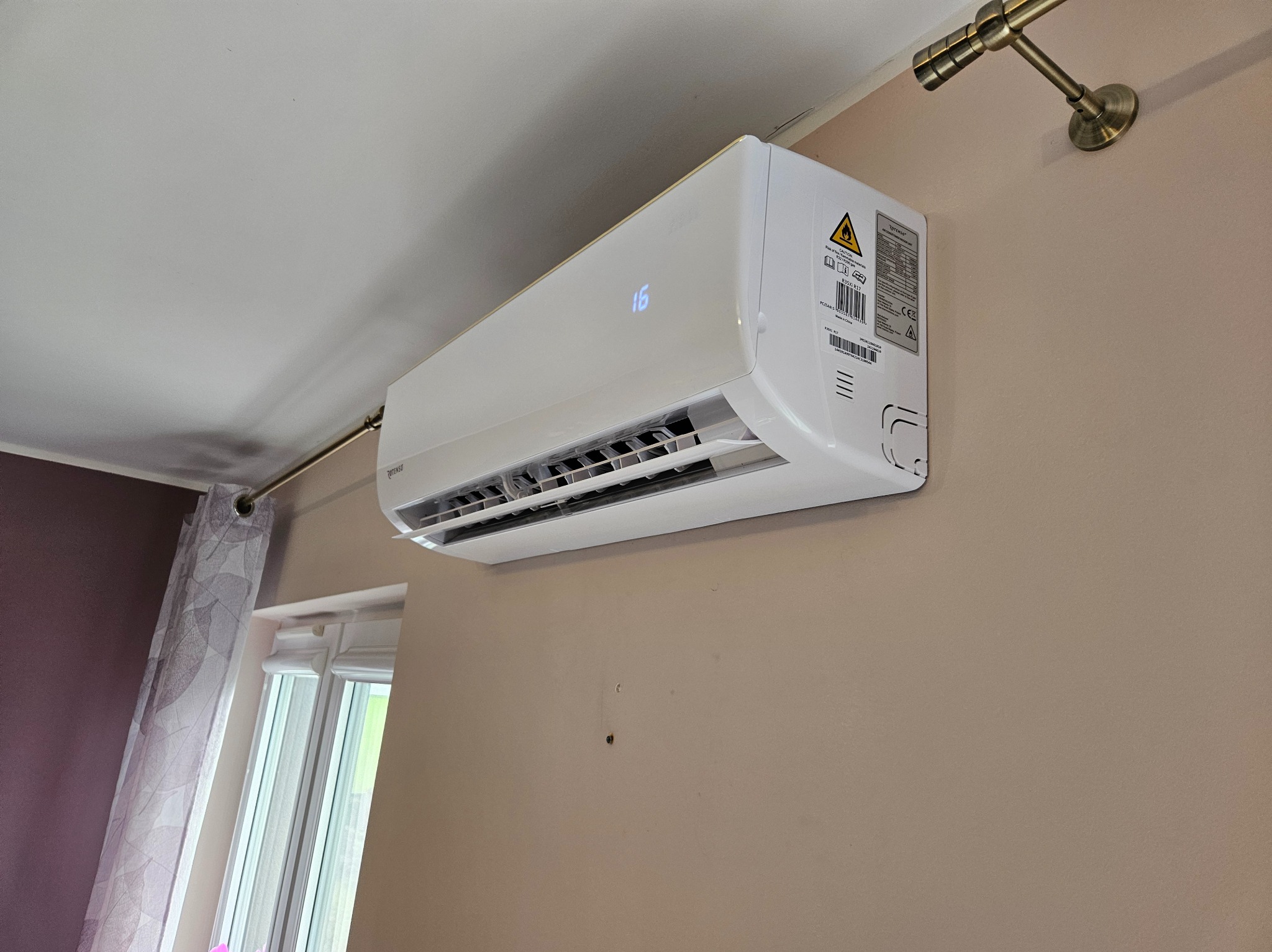 Biała klimatyzacja z wyświetlaczem temperatury (16°C) zamontowana na beżowej ścianie w pokoju z oknem i zasłoną w tle. Widoczny fragment karnisza.