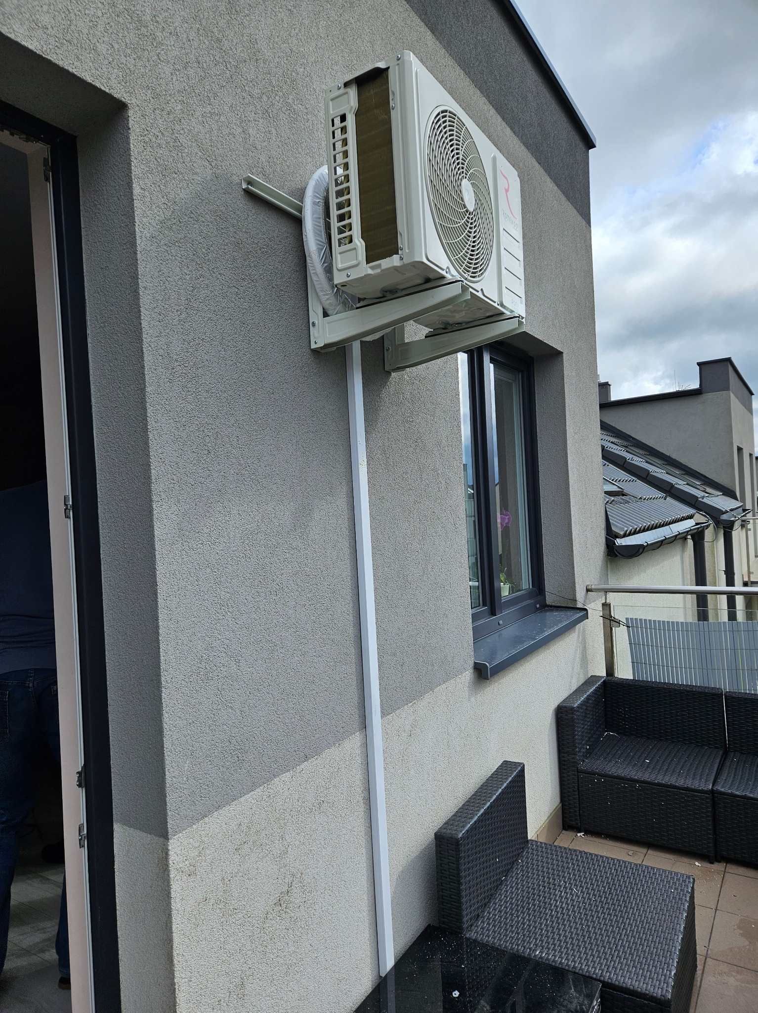 Zamontowana klimatyzacja ROTENSO na elewacji budynku z widocznymi elementami instalacji, rurami i uchwytami. Na balkonie meble ogrodowe z rattanu.