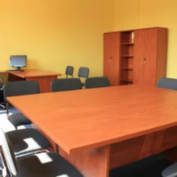 Sala konferencyjna z dużym drewnianym stołem, krzesłami z szarej tapicerki i żółtymi ścianami, widoczny komputer na biurku i szafa zamykana na klucz