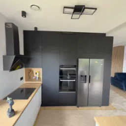 Nowoczesna, czarna kuchnia z drewnianym blatem i srebrną lodówką side-by-side. Na blacie wiertarka. Widok na salon z niebieską kanapą i drewnianą ścianką.