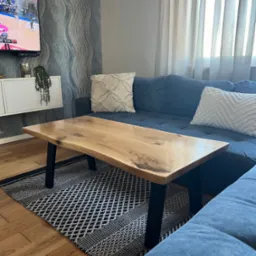 Stolik kawowy z blatem z surowego drewna na czarnych nogach, ustawiony na wzorzystym dywanie w salonie z niebieską kanapą i telewizorem w tle.