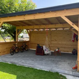 Drewniana pergola z czarnym dachem, oświetlona girlandą żarówek. Wewnątrz rowery, fotel i wiszący hamak. Tło: trawnik i drzewa. Widok z zewnątrz, słoneczny dzień.