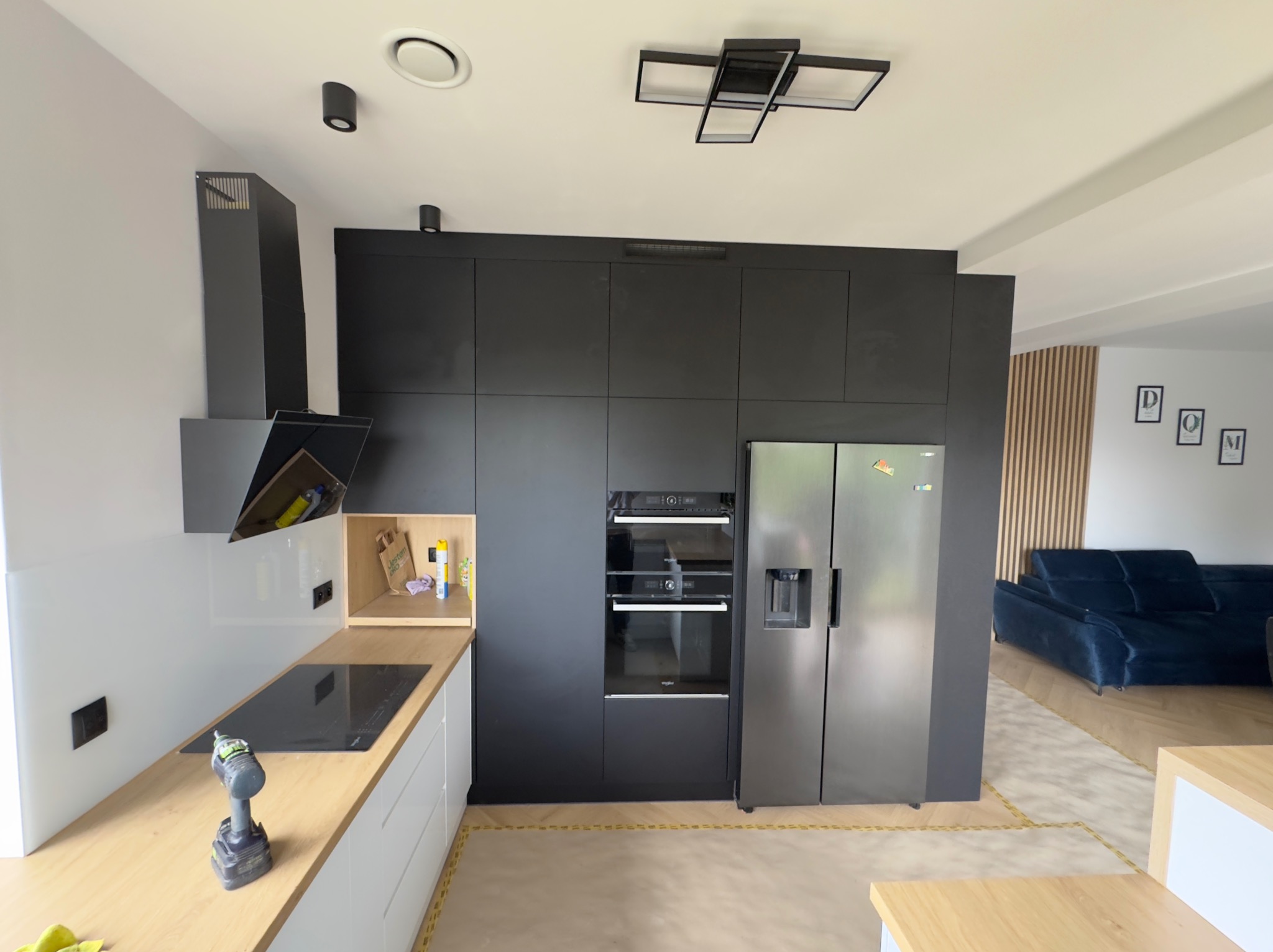 Nowoczesna, czarna kuchnia z drewnianym blatem i srebrną lodówką side-by-side. Na blacie wiertarka. Widok na salon z niebieską kanapą i drewnianą ścianką.