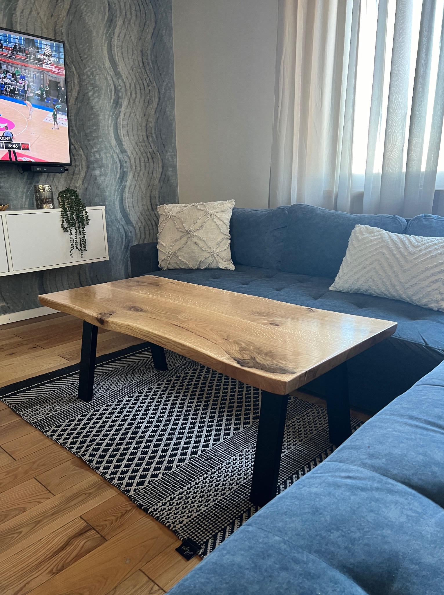 Stolik kawowy z blatem z surowego drewna na czarnych nogach, ustawiony na wzorzystym dywanie w salonie z niebieską kanapą i telewizorem w tle.
