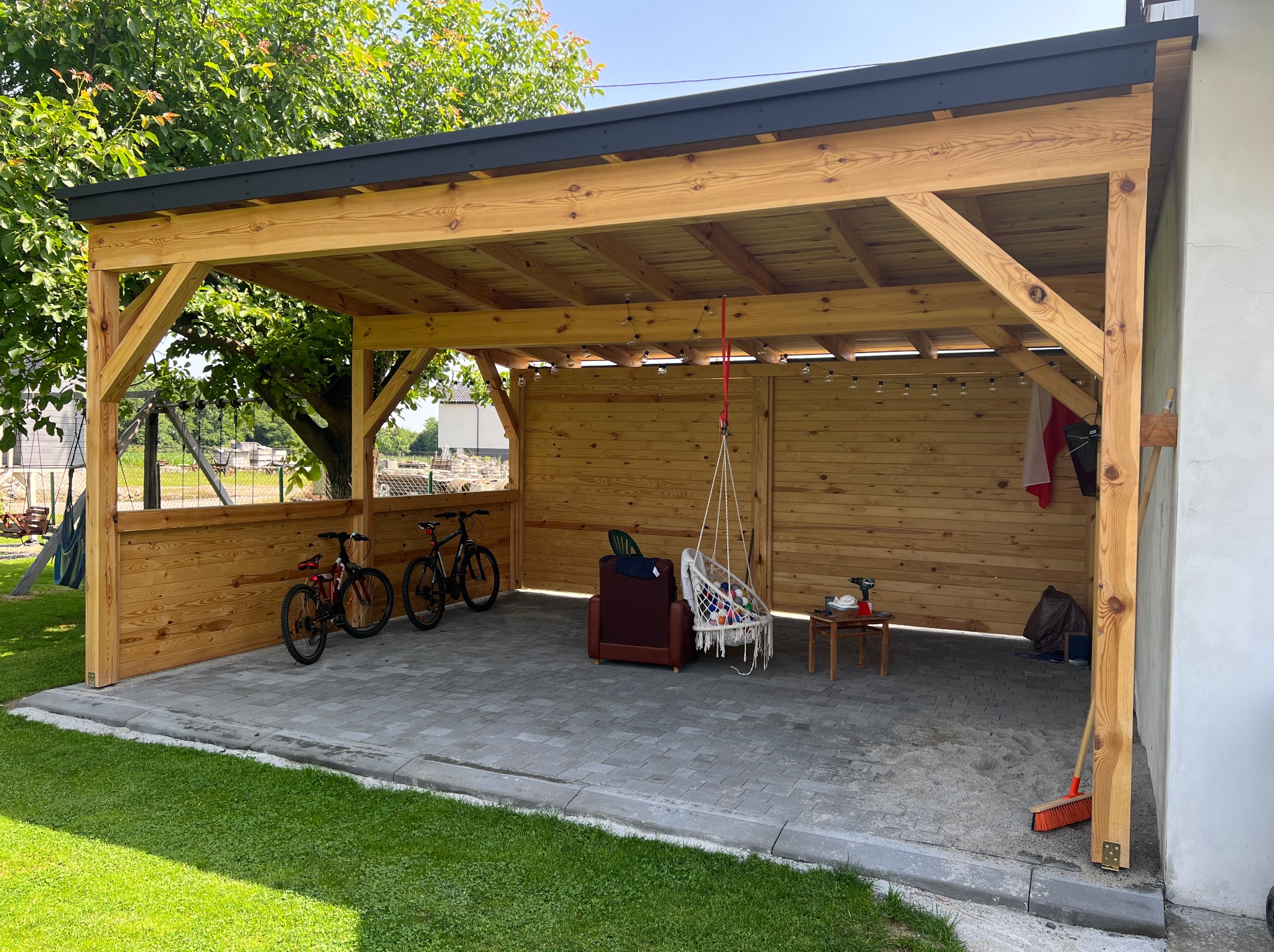 Drewniana pergola z czarnym dachem, oświetlona girlandą żarówek. Wewnątrz rowery, fotel i wiszący hamak. Tło: trawnik i drzewa. Widok z zewnątrz, słoneczny dzień.