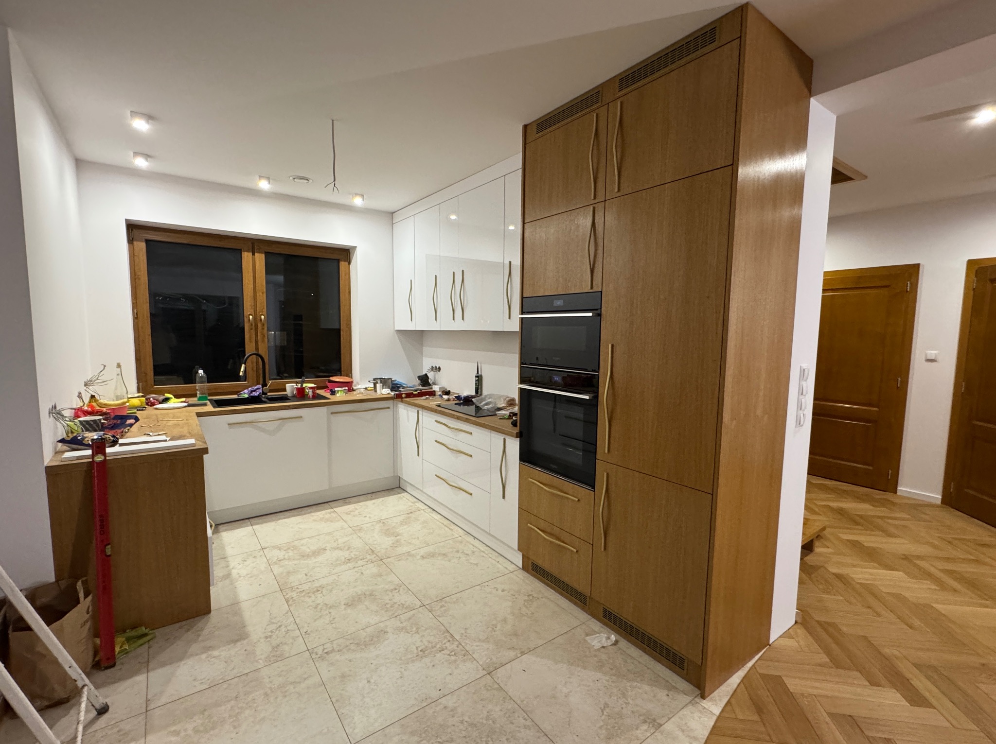 Nowoczesna kuchnia z białymi szafkami i drewnianymi akcentami, w trakcie montażu, widoczne narzędzia i poziomica, jasne płytki podłogowe, parkiet w jodełkę w tle.