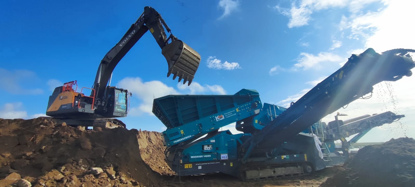 Ciężka koparka Volvo z podniesioną łyżką nad przesiewaczem mobilnym Powerscreen Warrior 1400X, pracującym na placu budowy pod błękitnym niebem z chmurami.