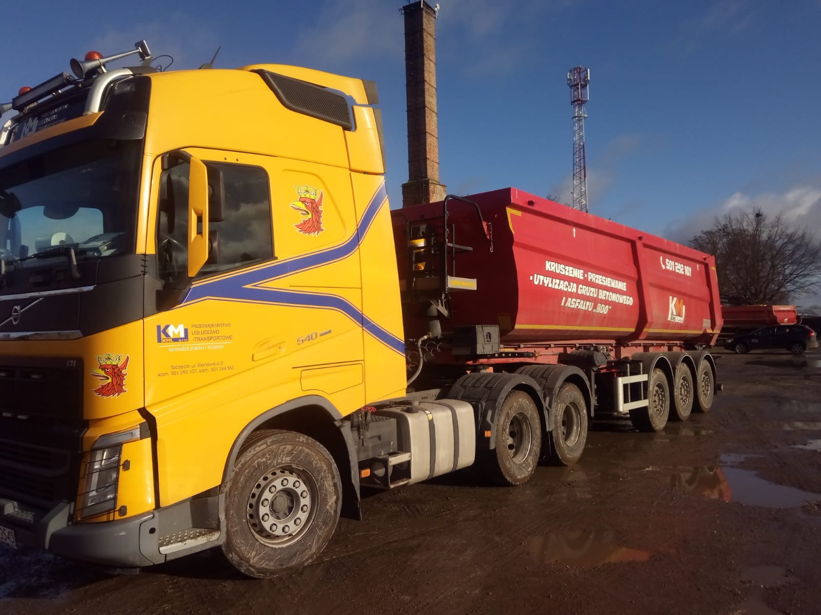 Żółta ciężarówka Volvo z czerwoną naczepą, na której widnieje napis 'Kruszenie - Przesiewanie, Utylizacja gruzu betonowego i asfaltu...', stoi na błotnistym terenie z kominem i wieżą...