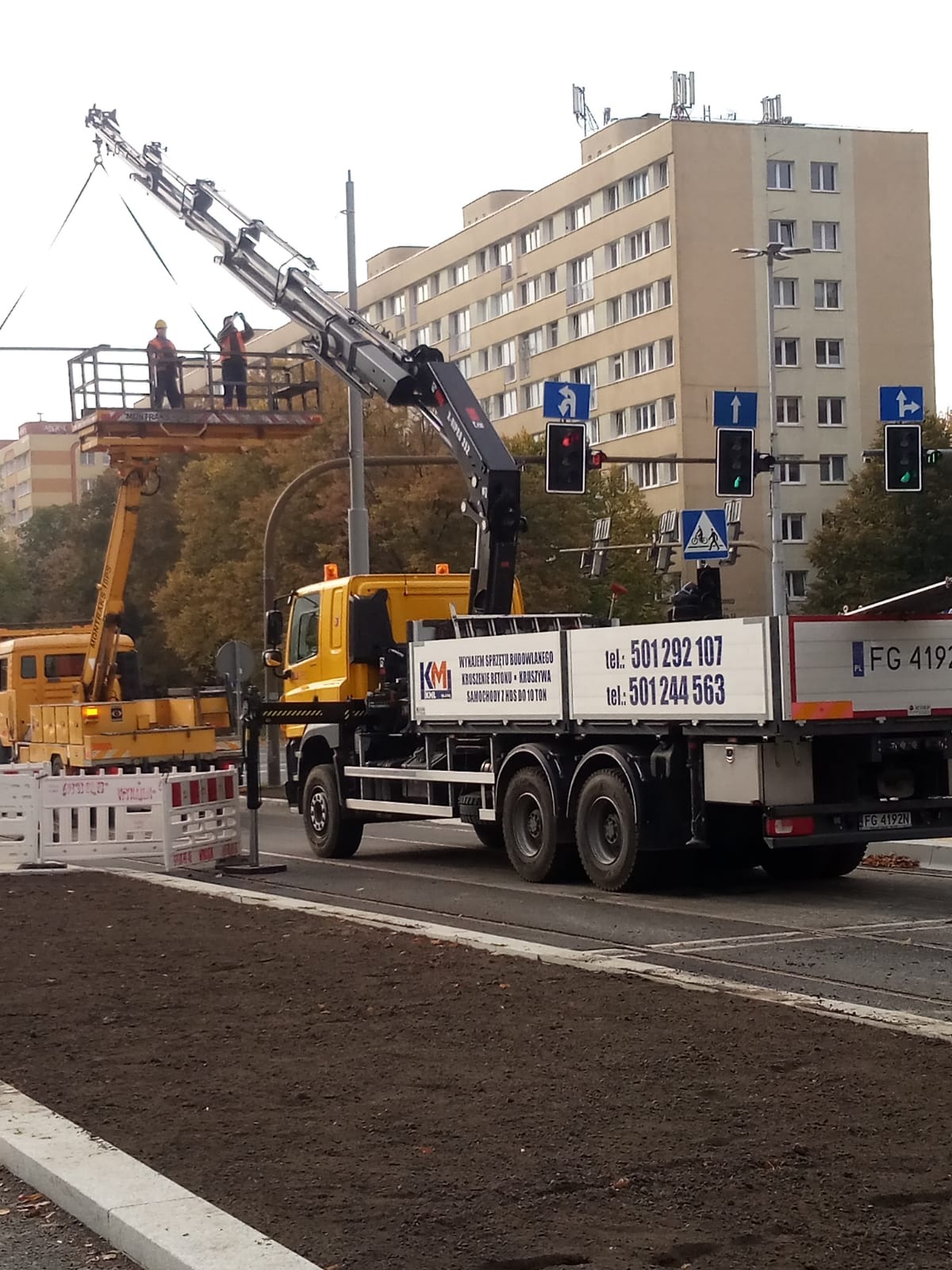 Żółty dźwig samochodowy z podnośnikiem koszowym i dwoma pracownikami na platformie, wykonujący prace przy instalacji nad ulicą w miejskiej scenerii.